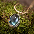 Load image into Gallery viewer, Et nærbillede af *Vedhæng - Eryn Design - Tranquill 14kt Guld* viser en stor, oval-formet grønlig-blå ædelsten sat i en indviklet sølvring. Vedhænget er lavet af Eryn Design og hænger på en flettet 14 karat guldkæde og hviler på en seng af mos. Ædelstenen reflekterer lyset og giver den et lysende udseende.