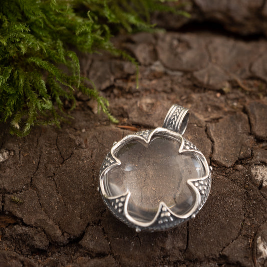 Her er en omformuleret version af sætningen ved hjælp af de givne produktdata:
Et gotlandsk krystalvedhæng formet som en blomst, sat i 925 sterling sølv. Den hviler på tekstureret træbark med grønt mos synligt i øverste venstre hjørne. Vedhæng Gotlandsk krystal Stor har en udsmykket, detaljeret metalindfatning, der minder om vikingetidens håndværk.