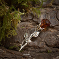 Load image into Gallery viewer, Broche Rose med Rav 925s Sølv, indviklet formet som en rose, sidder elegant på toppen af struktureret træbark. Dens kronblade er lavet af ægte rav, mens sarte sølvblade strækker sig yndefuldt ud fra stilken. Grønt mos omgiver brochen og forstærker den naturlige skønhed i denne romantiske scene. Et udsøgt stykke perfekt som en romantisk gav.