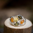 Load image into Gallery viewer, Sølvring Amber Drops 925s Sølv, med fem forskellige ædelsten arrangeret i et bølget, organisk mønster, vises på en træoverflade. Ædelstenene fremviser forskellige nuancer såsom rav, gul og grøn. Baggrunden er sløret for at holde fokus på det indviklede ringdesign.
