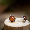 Load image into Gallery viewer, Et par Ørestikker Amber Key 925s Sølv, med runde rav-sten sat i sterlingsølv, hviler på en naturlig træoverflade. Den ene ørering står oprejst, mens den anden ligger ned og fremhæver det udsøgte håndværk og den rige, rødbrune nuance af ravstenene.