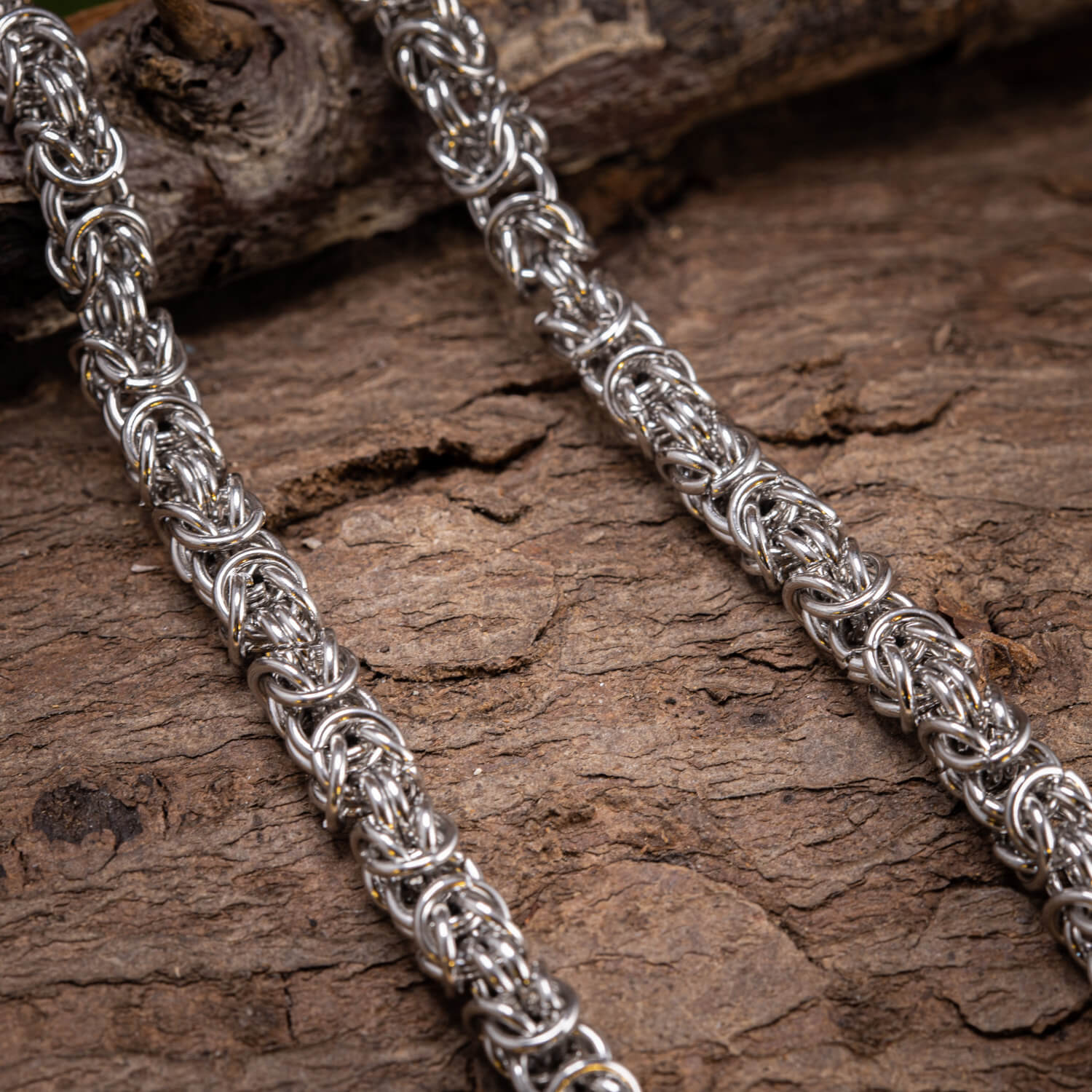 Et nærbillede af det indviklede Halskæde Kongekæde Stål 6 mm ringmaille smykker placeret på en ru, struktureret træoverflade. De sølvtonede metalled er sammenvævet i et komplekst, gentagende kongekædemønster, der viser detaljeret håndværk, der minder om vikingekunstneri.