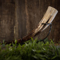 Load image into Gallery viewer, En poleret Drikkehorn Basic uden holder hviler vandret på et metalstativ. Hornet har nuancer af creme, brun og sort, sat mod en rustik træbaggrund. Grønt er synligt i forgrunden og tilføjer et strejf af natur, der minder om gammel vikingemarkeder.
