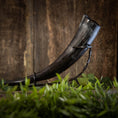 Load image into Gallery viewer, En elegant Drikkehorn Basic uden holder med en mørk nuance vises elegant på et sort metalstativ, sat mod en træbaggrund. I forgrunden introducerer sløret grønt løv et strejf af naturlig kontrast til den rustikke scene, hvilket fremkalder atmosfæren af traditionel vikingemarkeder.