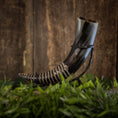Load image into Gallery viewer, En smukt udformet Drikkehorn-spiral hviler på et sort metalstativ, sat mod en rustik træbaggrund. Hornet har en skinnende, poleret finish med indviklede spiraludskæringer og er omgivet af frodigt grønt løv i bunden - perfekt til Vikingemarkeder eller som en elegant dekoration.