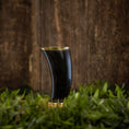 Load image into Gallery viewer, Et unikt Drikkeglas Horn (300 ml) fyldt med mørk væske står på en overflade prydet med levende grønt løv. Baggrunden har en rustik trætekstur, der skaber en naturlig og jordnær stemning.