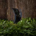 Load image into Gallery viewer, Drikkeglas Horn Plain (300 ml), et sort, buet glas, der ligner et poleret bøffelhorn, står oprejst på en overflade dækket af grønt løv. Baggrunden består af et struktureret, brunt træpanel.