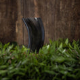 Load image into Gallery viewer, Drikkeglas Horn Plain (300 ml), et buet drikkehorn lavet af sort bøffelhorn, står oprejst bag en plet med grønt græs. Baggrunden har en rustik træoverflade, der forstærker scenens naturlige og jordagtige atmosfære.