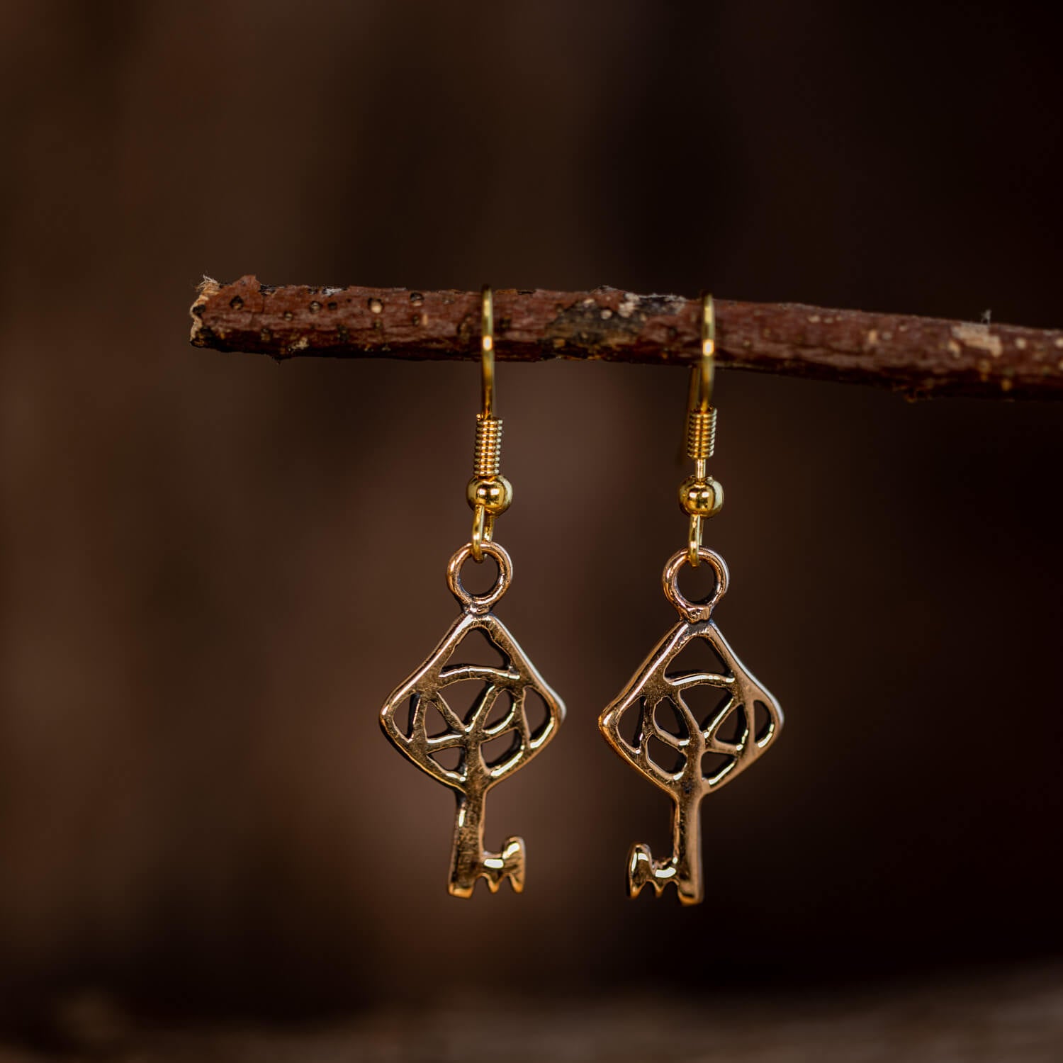 Et nærbillede af et par elegante Hængeøreringe Nøglen til Himmerriget Bronze hængende fra en gren. Øreringene, der er lavet i forgyldt sølv, har et geometrisk design, der ligner en nøgle med et indviklet trekantet og cirkulært mønster. Baggrunden er mørk og sløret, hvilket skaber en kontrast, der fremhæver de skinnende øreringe.