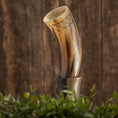 Load image into Gallery viewer, Drikkehorn Rune, prydet med indviklede mønstre, er placeret på et træstativ mod en rustik træbaggrund. I forgrunden tilføjer grønt græs og planter et naturligt præg til scenen og fremkalder stemningen af vikingemarkeder.