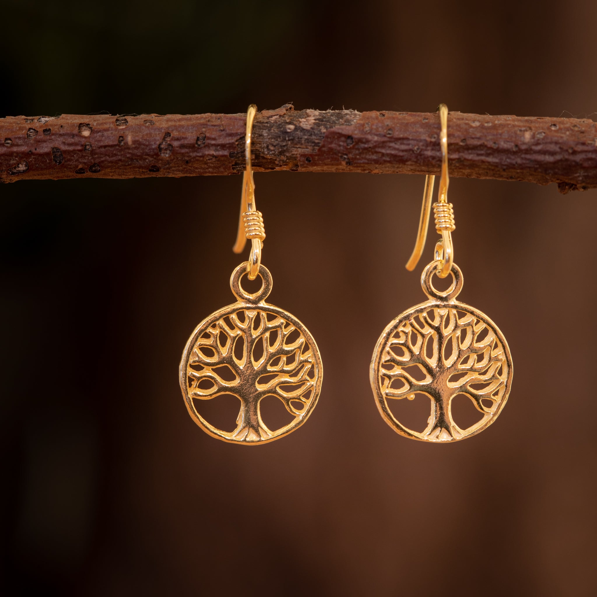 Et par Hængeøreringe Yggdrasil Livets Træ Minth Forgyldt 925s Sølv hænger fra en kvist. Det indviklede design viser et detaljeret livets træ inden for en cirkulær ramme. Den gyldne nuance står i kontrast til den slørede, mørke baggrund og fremhæver elegancen af smykket, der er lavet i 925'erne Sterling Sølv.