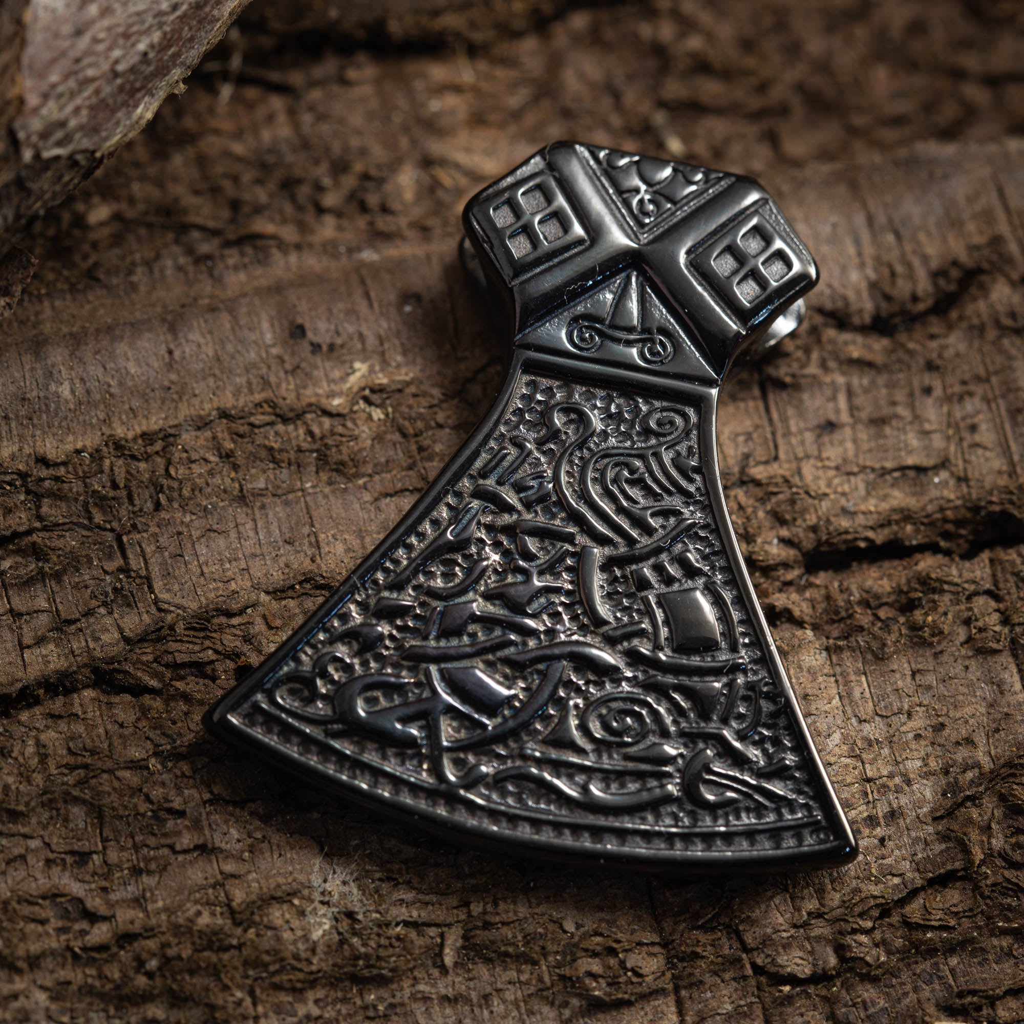 Et nærbillede af den indviklet designede Vikingeøkse Vedhæng Sort Stål fra BlaX Steel-serien hviler på en tekstureret, forvitret træoverflade, der viser detaljerede graveringer med mønstre og symboler, der minder om nordisk mytologi.