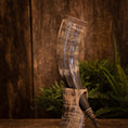 Load image into Gallery viewer, Drikkehorn Fenrir, lavet af indviklet designet bøffelhorn og poleret til en skinnende finish, vises oprejst ved hjælp af et udsmykket udskåret stativ. Baggrunden har rustikke træpaneler og levende grønne bregner, der giver scenen en naturlig og historisk følelse.
