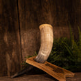 Load image into Gallery viewer, Et smukt udformet Drikkehorn Raw m. Thors Hammer Holder hviler i sit træstativ på en rustik træbaggrund, der minder om en gammel vikingeudstilling. Hornet viser en fantastisk gradient fra mørkt i bunden til lyst i spidsen, med grønt løv, der tilføjer et naturligt præg til baggrunden.