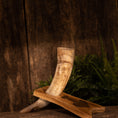 Load image into Gallery viewer, En rustik, tekstureret baggrund viser Drikkehorn Raw m. Thors Hammer Holder hvilende på et træstativ. Hornet viser en naturlig gradient af beige og brune toner, der minder om traditionel vikinge-dekoration. Grønt løv ses i baggrunden, hvilket tilføjer et jordisk præg til indstillingen.