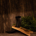 Load image into Gallery viewer, Drikkehornet Ak m. Thors Hammer Holder er udstillet mod en rustik trækulisse. Den hviler smukt på sit træstativ med antydninger af frodigt grønt løv i baggrunden, der tilføjer et naturligt touch til scenen. Denne opsætning er ideel til vikingemarkeder-entusiaster, der ønsker at ære traditioner.