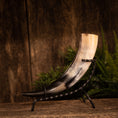 Load image into Gallery viewer, Drikkehorn Rune med metalholder er et poleret drikkehorn med en gradient, der går fra sort i spidsen til cremefarvet nær åbningen. Den er dekoreret med runegraveringer og fremvises på et elegant sort metalstativ mod en rustik træbaggrund, suppleret med grønt placeret på højre side.