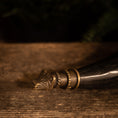 Load image into Gallery viewer, Et nærbillede af den dekorative ende af Drikkehorn Fenrir, med en udskåret metalspids formet som et dyrehoved. Hornet hviler på en rustik træoverflade, med baggrunden sløret og mørk, hvilket fremhæver spidsens indviklede dekoration.