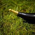 Load image into Gallery viewer, En poleret Drikkehornulv ligger på grønt græs. Det let buede, vintage sorte og guld horn har en skinnende sort krop og et guld ornamentalt mundstykke. Nærbilledet fremhæver græssets tekstur og hornets håndværk – perfekt til vikingemarkeder.