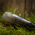 Load image into Gallery viewer, Drikkehorn Helleristning, prydet med indviklede keltiske udskæringer, ligger på græs mod en sløret træbaggrund, der fremkalder atmosfæren af vikingemarkeder.
