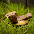 Load image into Gallery viewer, Et lille stykke solbrunt læder, der er lavet til en lille sandal, lægges på en træskive midt i levende grønt græs. Sandalen har et snøredesign, der viser detaljeret håndværk og en rustik, naturlig æstetik, perfekt til at tilføje et autentisk touch til din Drikkehorn Chief Dekoration.