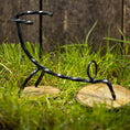 Load image into Gallery viewer, En Drikkehorn Spiral skulptur i sort metal står på to runde træbaser placeret på græs, med et plankeværk i baggrunden. Figuren, der minder om dekorationer på vikingemarkeder, ser ud til at være i en dynamisk, skæv positur.