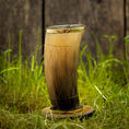 Load image into Gallery viewer, Et dekorativt Drikkeglas Horn med mønster (300 ml), med en gradient fra mørk til lysebrun og prydet med ætsede geometriske mønstre, vises på en rund træstand midt i grønt græs med et sløret træhegn i baggrunden.