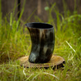 Load image into Gallery viewer, Et Drikkeglas Nature Horn (100 ml), lavet af flerfarvet bøffelhorn, hviler på en rund træbund midt i et græsareal. Baggrunden har et rustikt, sløret udseende, muligvis med træelementer.