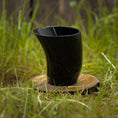 Load image into Gallery viewer, Drikkeglas Nature Horn (100 ml), et sort, buet drikkeglas lavet af bøffelhorn, står oprejst på en træskranke, omgivet af grønt græs med en sløret naturlig baggrund.