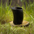 Load image into Gallery viewer, Et poleret Drikkeglas Nature Horn (100 ml) er placeret oprejst på en træskranke, sat på en plet med grønt græs udendørs. Det unikke glas tilføjer en rustik og jordnær følelse til scenen.