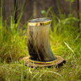 Load image into Gallery viewer, Et Drikkeglas Horn med Mønster (100ml) med indviklede udskæringer og en gylden rand er placeret oprejst på en træskive, omgivet af frodigt grønt græs. Rammerne fremstår rustikke og naturlige.