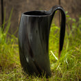 Load image into Gallery viewer, Et Vikingekrus Nature Horn (600-800 ml), med et slankt, mørkt, buet design med et håndtag, står på en plet græs. Kruset har en poleret, skinnende overflade og en organisk let snoet form. Baggrunden er lidt sløret og ser ud til at være af træ.