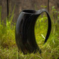 Load image into Gallery viewer, Et sort Vikingekrus Nature Horn (600-800ml) står på grønt græs med et rustikt plankeværk i baggrunden. Kruset, der efterligner den naturlige form af et dyrehorn, har en slank, blank finish og et stort buet håndtag.
