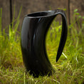 Load image into Gallery viewer, Et slankt, sort Vikingekrus Nature Horn (600-800ml) med sit unikke buede design står på græs med et plankeværk i baggrunden. Kruset har et utraditionelt håndtag, der passer problemfrit ind i dets moderne æstetik. De naturlige udendørs omgivelser står i smuk kontrast til den moderne stil på denne drikkekrus.