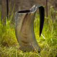 Load image into Gallery viewer, Et Vikingekrus Nature Horn (600-800ml) står oprejst på den græsklædte grund med et plankeværk i baggrunden. Hornet ligner et traditionelt vikinge-drikkekar og udviser naturlige variationer i farve, der spænder fra mørke til lysebrune nuancer.