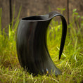Load image into Gallery viewer, Et Vikingekrus Nature Horn (600-800ml) med et sort, hornformet design og et buet håndtag sidder på grønt græs, sat mod en sløret træbaggrund. Dette krus efterligner et traditionelt drikkehorn, perfekt til dem, der værdsætter historien og æstetikken ved store drikkekrus.