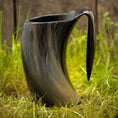 Load image into Gallery viewer, Et sort Vikingekrus Nature Horn (600-800ml) med en hornlignende form sidder på en græsklædt overflade. Denne unikke håndlavede krus har en blank finish, en bred åbning og et buet håndtag, der ligner et horn. Baggrunden er sløret, hvilket understreger kruset som fokus.