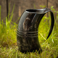 Load image into Gallery viewer, Et poleret sort Vikingekrus Helleristning Horn (600-800ml) med to dekorative bånd vises oprejst på græs med en træbaggrund. Håndlavede hornkrus har et håndtag, der giver den et kruslignende udseende.