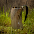 Load image into Gallery viewer, Et poleret, hornformet Vikingekrus Helleristning Horn (600-800ml) med et håndtag sidder på frodigt grønt græs. Baggrunden er en rustik trævæg, der forstærker scenens naturlige og historiske fornemmelse. Håndlavede hornkrus har subtile linjer og varierende nuancer af brun, tan og sort.