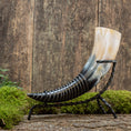 Load image into Gallery viewer, Drikkehorn-hjorten, der har et poleret horn med en gradient fra sort i bunden til beige i spidsen, er elegant monteret på et sort smedejernsstativ og hviler på en mosbeklædt træoverflade mod en rustik træbaggrund - ideel til vikingemarkeder.