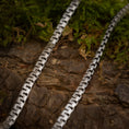 Load image into Gallery viewer, Nærbillede af to Halskæde Panzering 5,5 mm 925s Sølv-kæder lagt parallelt på et stykke struktureret, barkbeklædt træ. Grønt mos omgiver halskæden og tilføjer et naturligt element til scenen. Kæderne har et zigzag-mønster og et geometrisk, sammenhængende design i kontrast til den organiske baggrund.