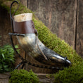 Load image into Gallery viewer, Et raffineret Drikkehorn Raw med Brun Læderstrop, elegant præsenteret på et snoet metalstativ. Baggrunden viser en rustik træoverflade prydet med grønt mos og løv, der forstærker den naturlige æstetik og fremkalder charmen ved vikingemarker.
