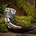 Load image into Gallery viewer, Drikkehorn Med Sort Læderholder, et poleret og buet bøffelhorn med sort og hvid marmorering, er sikkert fastgjort med en læderrem og snøre. Den hviler på en bund af grønt mos mod en rustik træbaggrund, der fremkalder vikingemarkedernes naturlige og jordnære stemning.