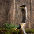 Load image into Gallery viewer, Produktbeskrivelse: Drikkehorn Ordained Raw, et rustikt bøffelhorn drikkehorn med metalkant, er smukt udstillet på en træbaggrund. Den står oprejst på grønt mos, med en lille plante indlejret bagved. Hornets overflade fremviser naturlige mørke og lyse mønstre, der fremkalder charmen fra Vikingemarkeder og tilføjer autenticitet til ethvert Roskilde showroom.