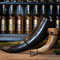 Load image into Gallery viewer, To udsøgt udformede drikkehorn, inklusive Drikkehorn Giant 1,5 liter, i forskellige størrelser og farver vises på et træbord. Hver bøffelhorn hviler på et metalstativ, og en række flasker er synlige i baggrunden på hylder. Rammerne er rustikke og elegante, der minder om traditionel vikingemarkeder.