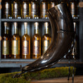 Load image into Gallery viewer, En Drikkehorn Giant 1,5 liter står udstillet på et stativ foran en hylde fyldt med forskellige flasker spiritus. Det buede og skinnende stykke er fremhævet med en strimmel grønt mos i bunden, der minder om skatte set på vikingemarkeder.