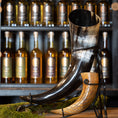 Load image into Gallery viewer, En udstilling af to drikkehorn, det ene med det imponerende Drikkehorn Giant 1,5 liter i sort bøffelhorn og det andet mindre og brunt, sat på et stativ med en mosbeklædt bund. Baggrunden byder på hylder fyldt med forskellige drikkevarer på flaske, der minder om vikingemarkeder.
