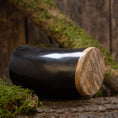 Load image into Gallery viewer, En poleret sort Drikkeglas Horn Plain (500ml) med en glat træbund hviler på en rustik overflade omgivet af grønt og træbark. Baggrunden af træplanker fremhæver den naturlige og jordiske stemning.