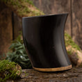 Load image into Gallery viewer, Drikkeglas Horn Plain (500 ml) står elegant oprejst på en tekstureret træoverflade og viser dets glatte, buede sorte bøffelhorn med en solbrun base. Baggrunden inkluderer træstykker og pletter af grønt mos, der bidrager til scenens rustikke og naturlige stemning.