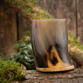 Load image into Gallery viewer, Et poleret Drikkeglas Horn Plain (500 ml) med et buet design og træbund hviler på en mosbeklædt træstamme. Baggrunden viser en rustik træoverflade, delvist prydet med grønt mos og løv.